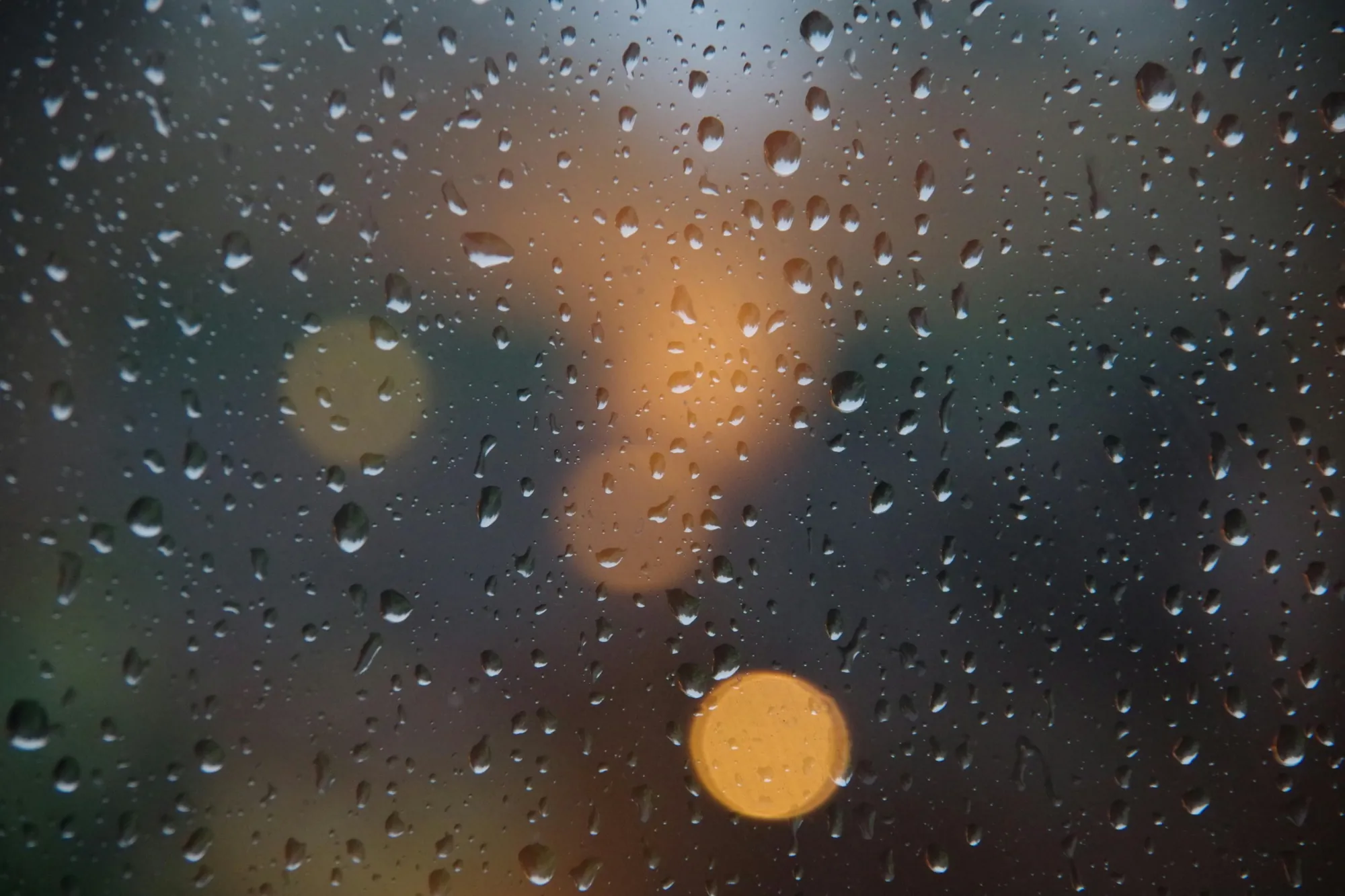 Raindrops on fogged glass with soft, out-of-focus lights behind, suggesting a quiet moment of reflection