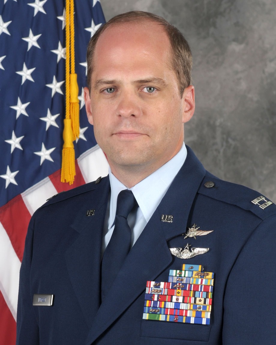 Todd Temaat in a formal Air Force portrait: dress blues with captain’s bars, aircrew wings and rows of service ribbons, a US flag behind him