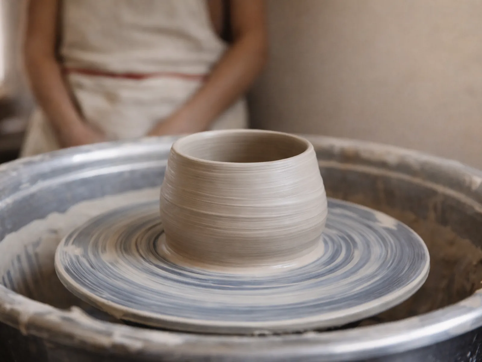 An unfinished clay pot on a potter’s wheel, a person in an apron out of focus behind it with their hands away from the clay